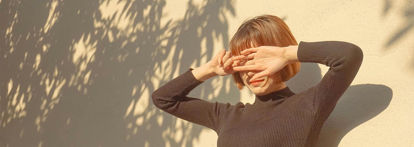 Woman shielding her eyes from the sun while standing against a textured exterior wall with leafy shadows.