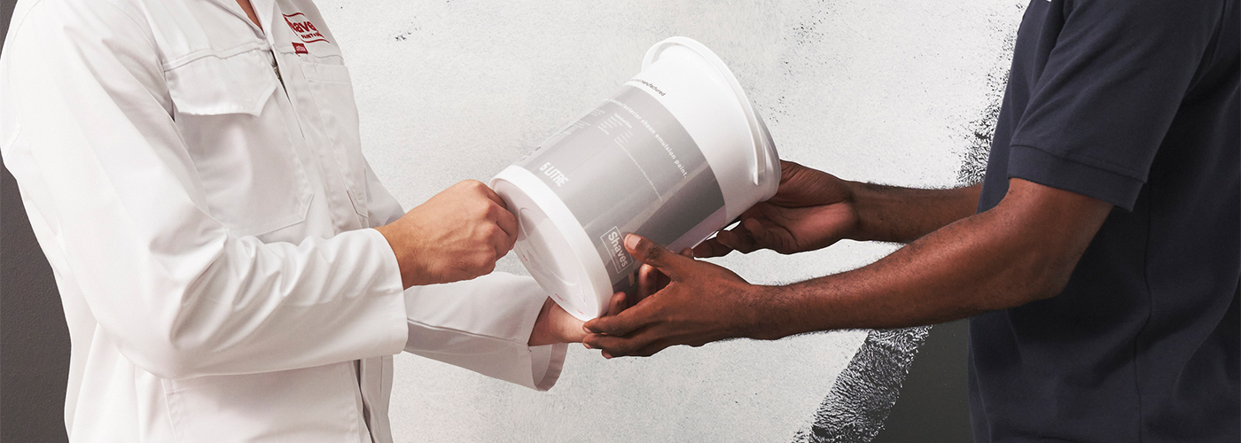 Two people exchanging a paint bucket against a partially painted grey and white wall.
