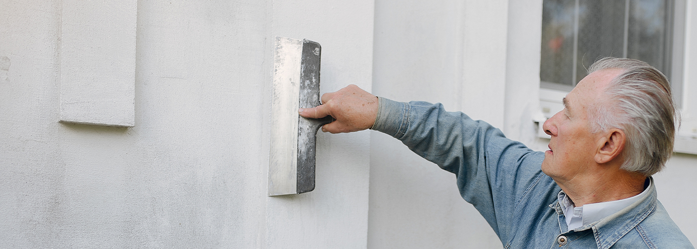 A man in a blue work shirt applies plaster or skim coat to an exterior white wall using a large scraper.