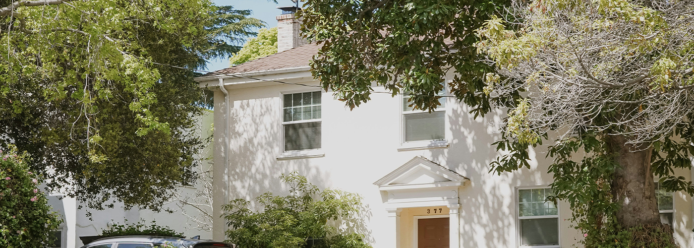 A beige two-storey house with white trim and a tree casting shadows on the front lawn.