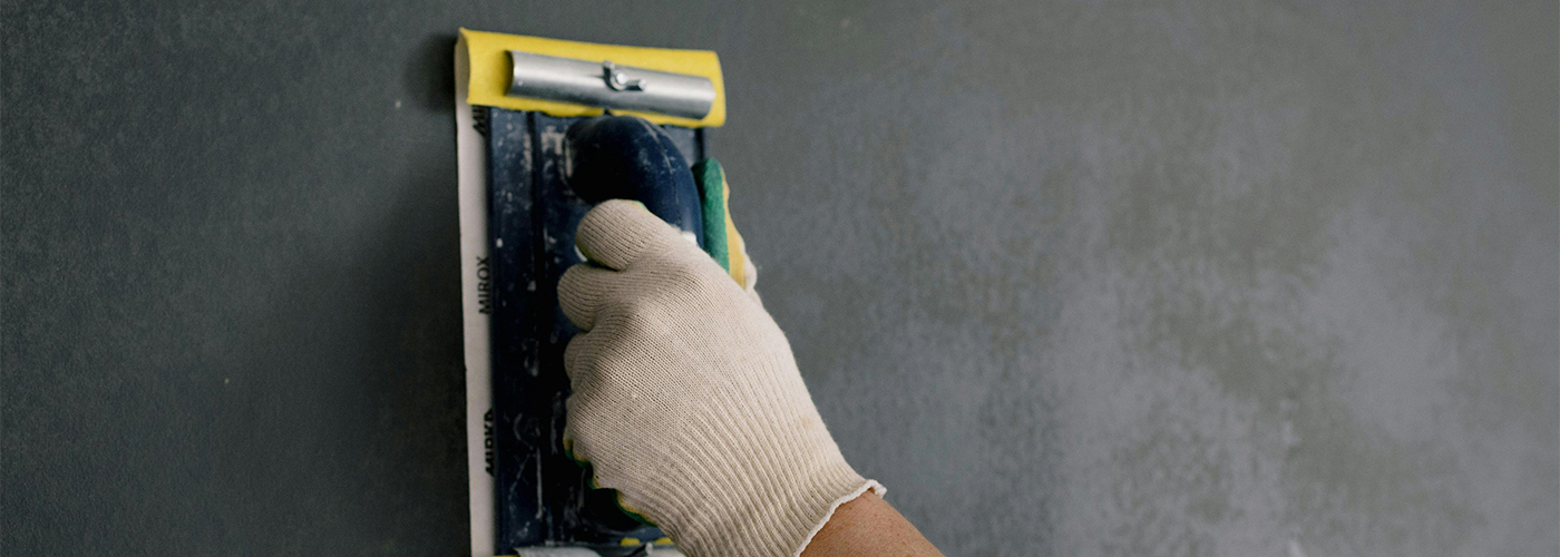 A gloved hand smoothing a dark grey wall with a flat trowel tool during surface preparation.